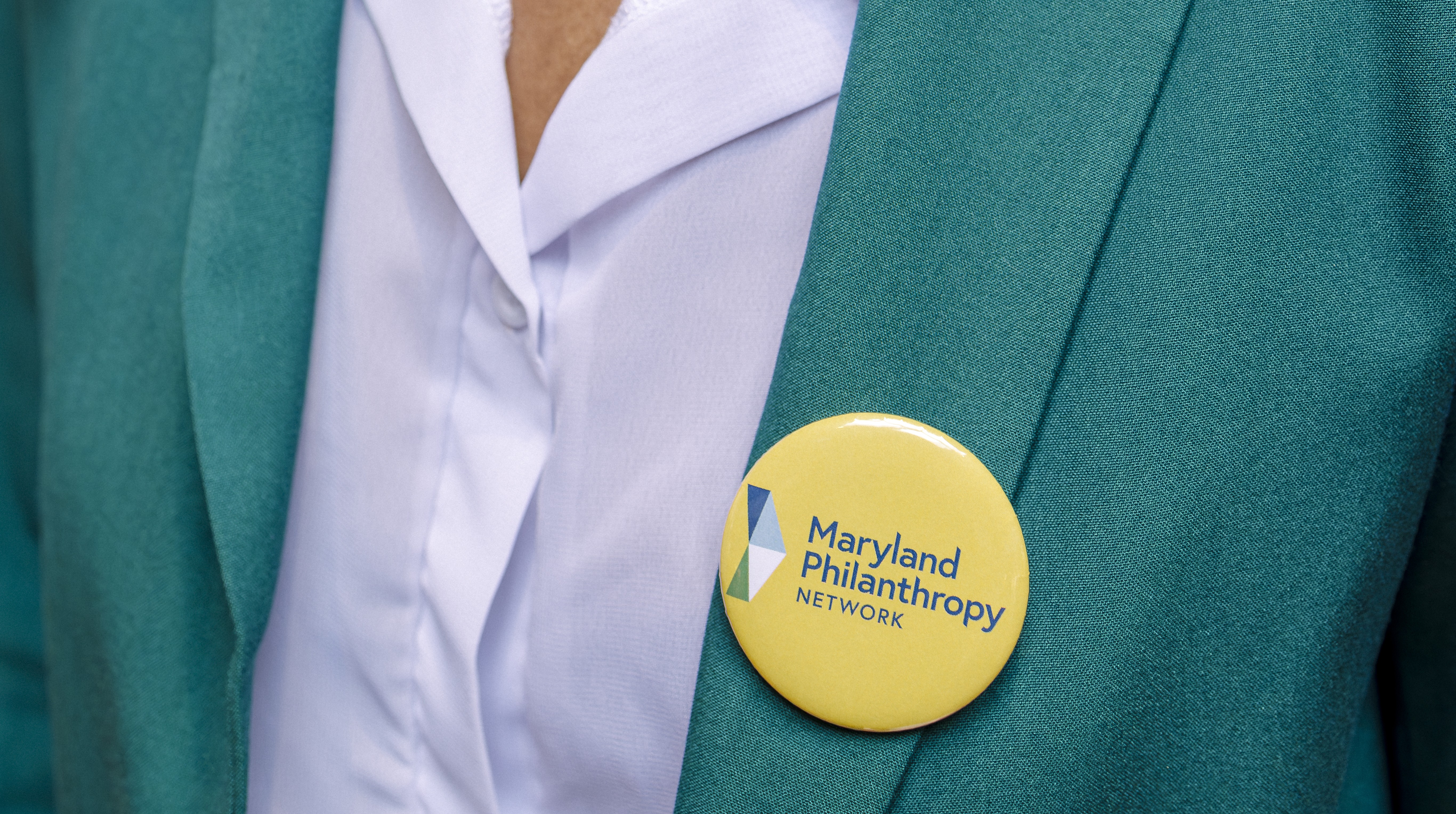 A yellow pin-on button with MPN's logo adorns someone's teal blazer.