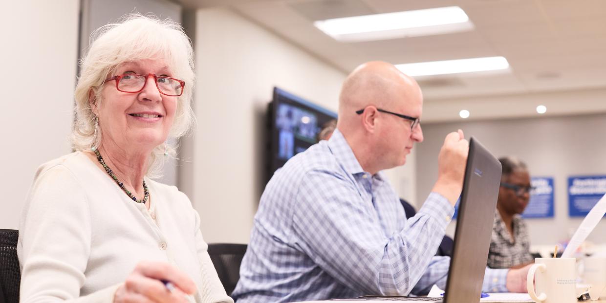 Photo of two people seated at the conference room tables with laptops in front of them.