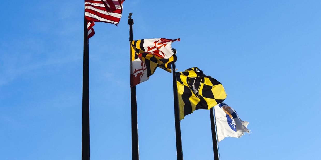 Four flags, including the USA and Maryland State flags, fly in the wind on a blue sky.