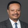 Headshot of a Black man with short hair and facial hair in a dark grey suit, white collared shirt, and blue tie.